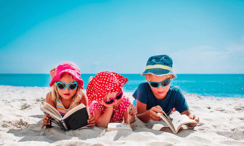 Three kids lying on tummies on beach, reading books