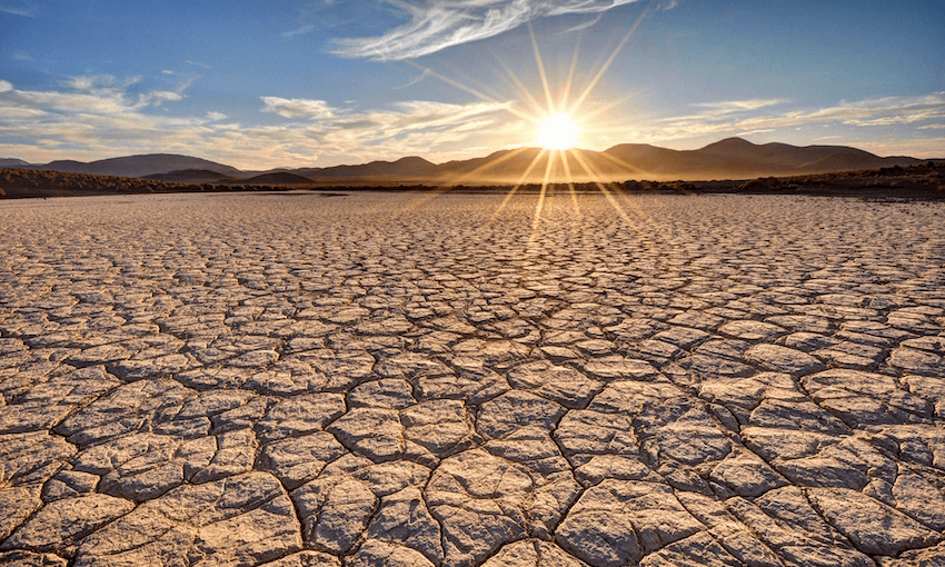 Photo of dry, cracked earth in the desert with a sunset over a mountain range in the background.