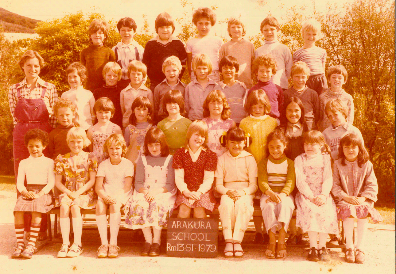 A sepia-toned class photo of Arakura School, Room 13, from the year 1979. The group consists of young students and a teacher, all posing outdoors with a sign displaying the school and class information. The background includes bushes and a hillside.