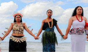 Three women stand hand in hand at the water’s edge against a backdrop of blue sky and clouds. Each wears traditional attire with vibrant patterns, natural materials, and accessories, reflecting their cultural heritage.